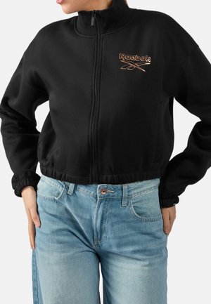 Frau trägt eine schwarze, kurze Reebok-Jacke mit Reißverschluss und hellblaue hochtaillierte Jeans, die Hände ruhen auf den Hüften.