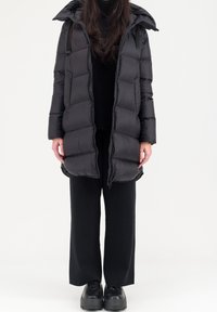 Zwarte pufferjas met een capuchon, voorzien van een gewatteerd ontwerp, ritsluiting en oversized silhouet. Draagt over zwarte kleding en stevige schoenen.