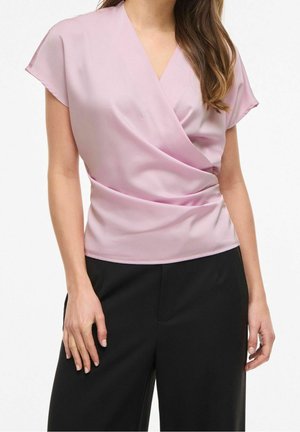 Femme portant un chemisier cache-cœur à manches courtes rose clair associé à un pantalon noir taille haute sur un fond uni.