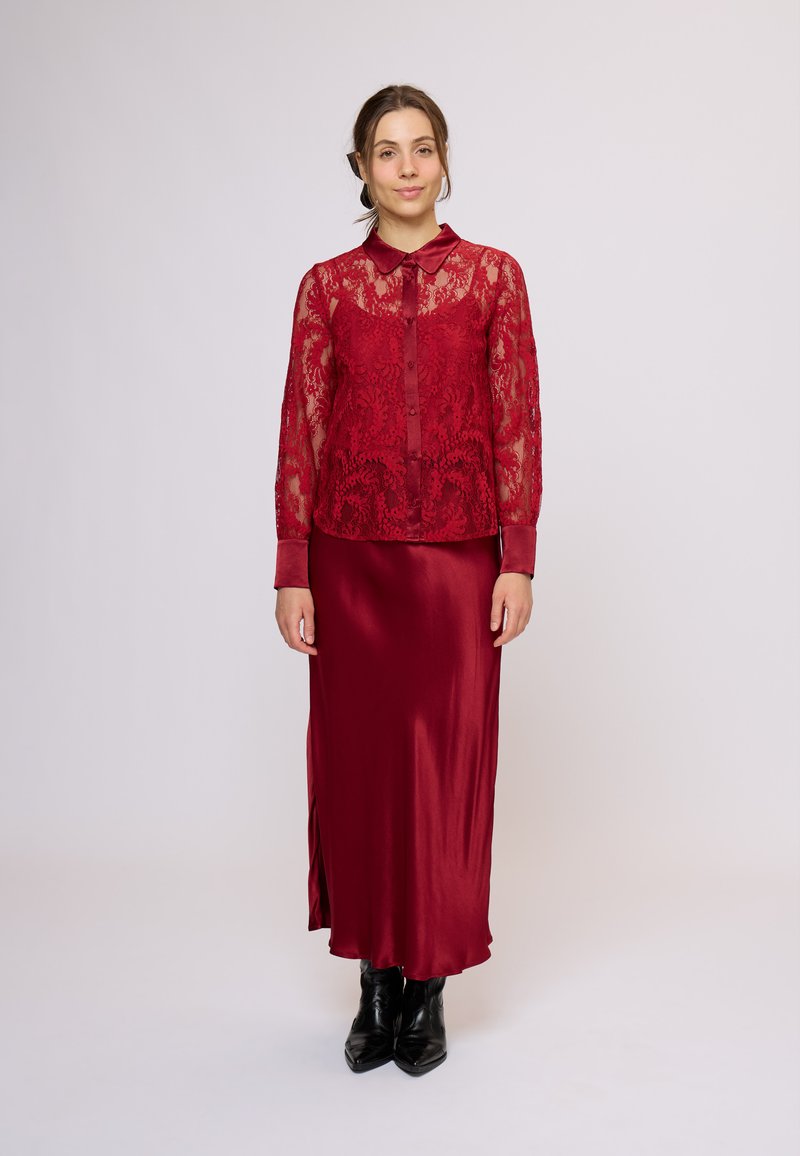 Cămașă cu nasturi din dantelă roșie, cu mâneci lungi, asortată cu o fustă lungă din satin, cu o despicătură laterală și cizme negre până la gleznă.