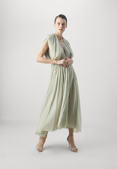 Rochie lungă de culoare verde deschis, cu material drapat, decolteu adânc în V și mâneci în formă de capă. Materialul texturat se scurge până la glezne. Tocuri deschise la culoare.
