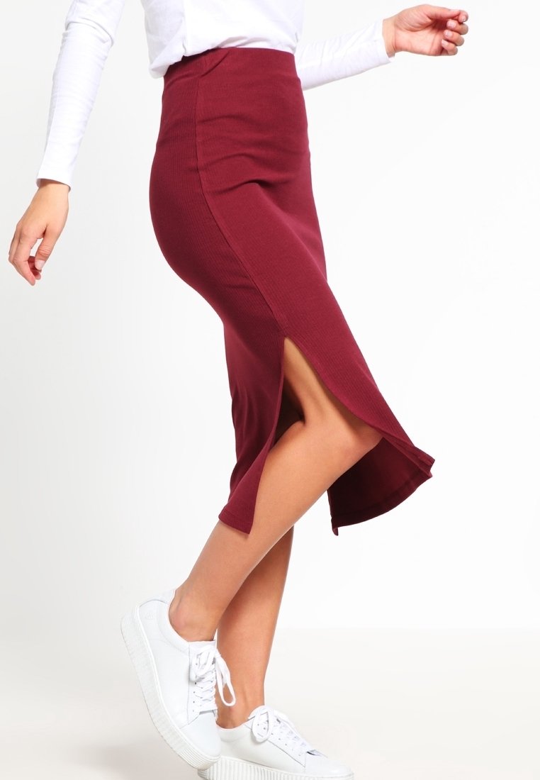 Femme portant une jupe midi bordeaux avec une fente latérale et des baskets blanches, vue du milieu du torse jusqu’aux pieds sur un fond uni.