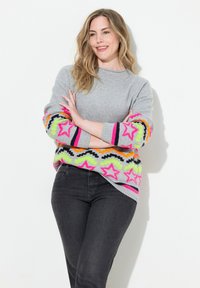 Grå tröja med flerfärgade stjärnmönster på ärmarna, med rosa, orange och gröna accenter. Bärs med mörka jeans.