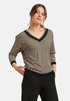 Zwart en beige gestreepte gebreide trui met V-hals, voorzien van een chevronpatroon, ribgebreide mouwen en zoom. Model poseert met handen in de zakken.