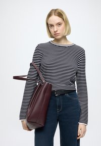 Grand sac cabas en cuir bordeaux avec une finition texturée et une forme souple, porté par une personne portant un haut à manches longues rayé noir et blanc.