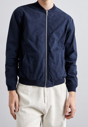 Homme portant une veste bomber bleu marine zippée avec poignets et ceinture côtelés, associée à un pantalon en velours côtelé écru.
