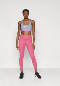 Μωβ αθλητικό σουτιέν με το λογότυπο "NIKE PRO", συνδυασμένο με ροζ εφαρμοστά κολάν με υφή. Μαύρα αθλητικά παπούτσια ολοκληρώνουν την εμφάνιση.