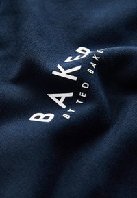 Navy blue fabric with raised white "BAKED by Ted Baker" text on a slightly wrinkled surface.