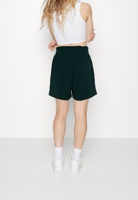 Žena s dlouhými vlnitými vlasy má na sobě bílý bezrukávový crop top, tmavě zelené šortky s vysokým pasem, bílé ponožky a bílé tenisky, stojí se zkříženýma rukama.