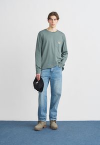 Long-sleeve green shirt with a chest pocket, paired with light blue jeans and beige footwear. Black cap held in left hand.
