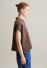 Braun kurzärmliges Poloshirt mit Kragen, locker sitzend und mit geradem Saum. Glatte Textur und einfaches Knopfdetail am Ausschnitt.