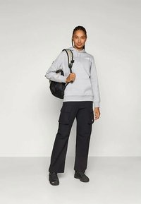 Femeie îmbrăcată într-un hanorac gri cu glugă, pantaloni cargo negri, adidași negri și purtând un rucsac negru pe un umăr.