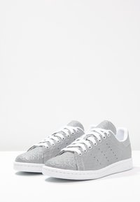 Zapatillas texturizadas grises con cordones blancos, que presentan un patrón escamoso en la parte superior, suela blanca y detalles perforados en los laterales.