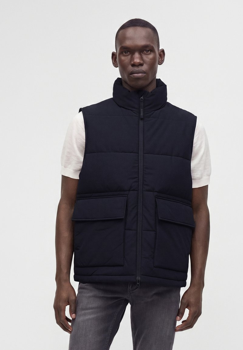 Άνδρας που φοράει μαύρο αμάνικο μπουφάν με μεγάλες τσέπες μπροστά πάνω από λευκό κοντομάνικο πουκάμισο και σκούρα γκρι τζιν, στέκεται μπροστά σε απλό φόντο.