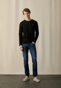 Chemise noire à manches longues avec un petit logo sur la poitrine, associée à un jean bleu et des baskets beiges, sur un fond neutre.