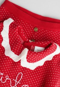 Roter Strickpullover mit einem gewelltem weißen Kragen, der kleine weiße Herzen und goldene Knöpfe auf dem Rücken hat. Markenlabel sichtbar.