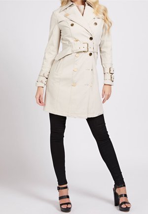Kvinde iført beige trenchcoat med bælte og guldknapper, sorte leggings og sorte sandaler med høje hæle står foran en ensartet baggrund.