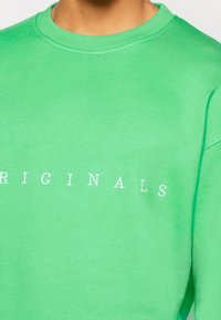 Grön bomullströja med rund halsringning, prydd med vit text "ORIGINALS" över bröstet. Slät textur, avslappnad passform.