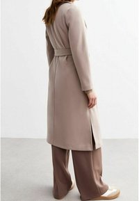 Person klädd i en lång beige kappa med bälte, vida bruna byxor och vita sneakers, står sidledes mot en ljusgrå golv- och vit väggbakgrund.