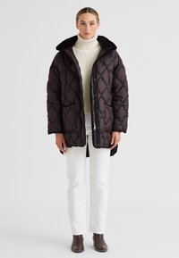 Frau in einem dunkelbraunen, gepolsterten Mantel mit Fellkragen über einem cremefarbenen Rollkragenpullover, weißen Hosen und braunen Stiefeletten, vor einem weißen Hintergrund stehend.