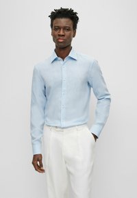 Camicia azzurra chiara con una leggera texture, maniche lunghe e colletto classico, abbinata a pantaloni bianchi.