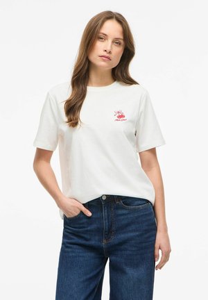 Jeune femme aux longs cheveux bruns portant un t-shirt blanc avec un petit motif de cerise rouge et un jean bleu, main dans la poche, fond neutre.