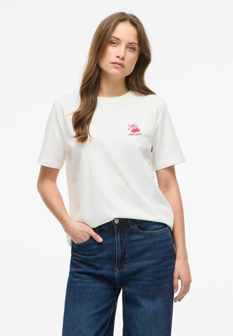 Jeune femme aux longs cheveux bruns portant un t-shirt blanc avec un petit motif de cerise rouge et un jean bleu, main dans la poche, fond neutre.