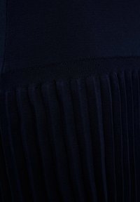 Tummansininen laskostettu kangas, jonka yläosa on sileä, ja sauma on varustettu kontrastivärisellä ribbitekstuuriin. Keskittyminen kankaisiin ja väriin.