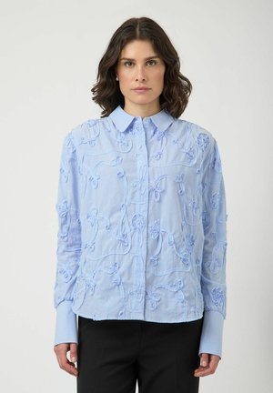 Kvinde iført en lyseblå skjorte med blomsterbroderi og forlængede manchetter, kombineret med sorte bukser, stående foran en ensfarvet baggrund.