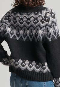 Zwarte gebreide trui met witte en grijze geometrische patronen over de schouders, mouwen en taille, gedragen met blauwe spijkerbroek.