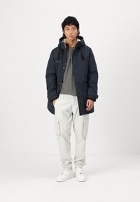 Abrigo acolchado de un azul marino oscuro con capucha y cordones, llevado sobre una camiseta gris y pantalones cargo claros. Zapatillas blancas completan el atuendo.