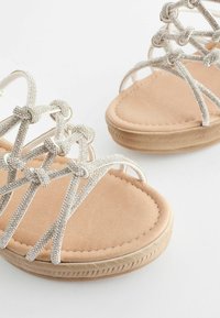 Des sandales argentées à lanières présentent une semelle intérieure beige douce, des détails de nœud décoratifs et une semelle en caoutchouc texturée pour une meilleure adhérence.