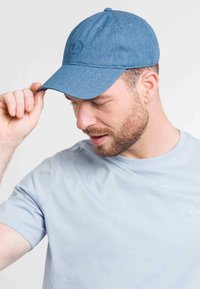 Denim mėlyna kepurė su išlenktu vizoriumi, turinti subtilius siuvinėjimus ir ventiliacines skyles. Derinama su šviesiai mėlyna trumpomis rankovėmis marškiniais.