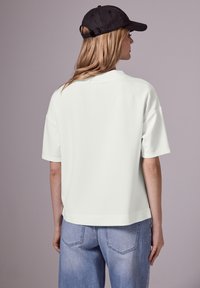 Camisa polo blanca de manga corta con corte recto, que presenta un collar plano y una textura sutil, combinada con unos vaqueros azul claro y una gorra negra.