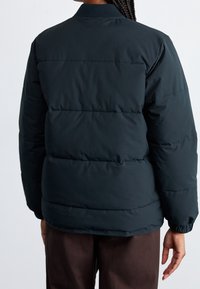 Giubbotto imbottito verde scuro con colletto a coste. Presenta sezioni trapuntate e polsini elastici. Texture liscia e opaca, con vestibilità normal.