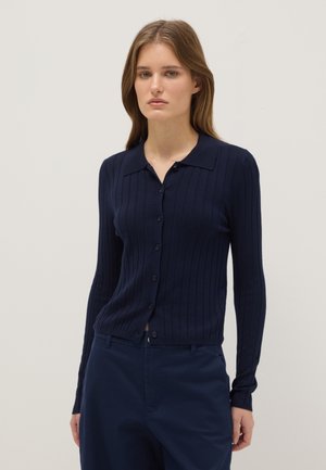 Woman wearing a navy blue ribbed button-up cardigan with a collar and matching navy trousers against a plain light background.