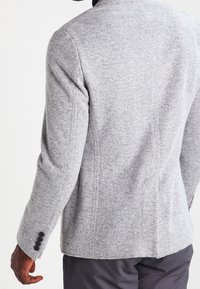 Hombre vistiendo un blazer de lana gris claro con botones negros en el puño, mostrado de espaldas y de lado, combinado con pantalones oscuros.