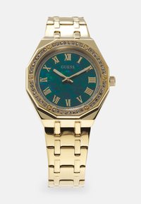 Gouden octagonale horloge met een teal iriserende wijzerplaat, Romeinse cijfers, kristallen accenten rond de bezel en een gepolijste metalen schakelarmband.