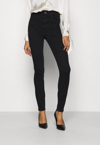 Jeans ajustados negros hechos de denim elástico, con una cintura alta, cierre de botón y una textura suave. Combinados con tacones de punta negra.