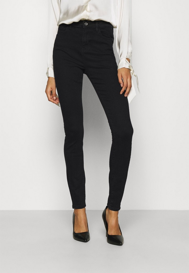 Jeans ajustados negros hechos de denim elástico, con una cintura alta, cierre de botón y una textura suave. Combinados con tacones de punta negra.