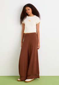 Top corto in cotone a coste color crema con bordi gialli a volant, abbinato a pantaloni brown a gamba larga con dettagli in pizzo all'orlo e scarpe bianche.