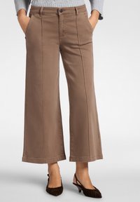 Väljä lahkeinen, ruskeasta kankaasta valmistetut housut etutaskuilla ja sileällä pinnalla. Sisältää napin sulkemisen ja näkyvät saumat lahkeiden varrella.