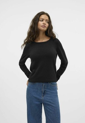 Zwarte, gestructureerde lange mouw top met een wijde halslijn, gecombineerd met hoog getailleerde blauwe jeans. De stof heeft een subtiel geometrisch patroon.