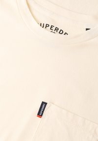 Camiseta color crema hecha de algodón, con cuello redondo y un bolsillo en el pecho que presenta un diseño de etiqueta con acentos en azul marino, rojo y blanco.