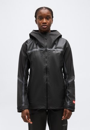 Žena oblečená v čiernej nepremokavej bunde Columbia Titanium s kapucňou a v čiernych nohaviciach, stojaca pred jednoduchým bielym pozadím.