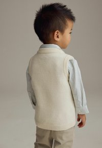 Gilet en tricot crème avec un col à côtes, porté par-dessus une chemise rayée bleu clair. L'enfant a les cheveux noirs et est positionné de dos, face au spectateur.