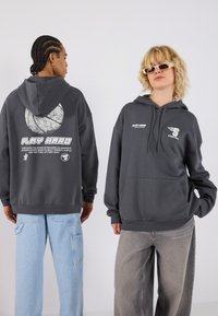 Grå oversized hettegenser med frontlomme, snorhette og trykte grafikker; kombinert med lys blå og grå vidbukser.