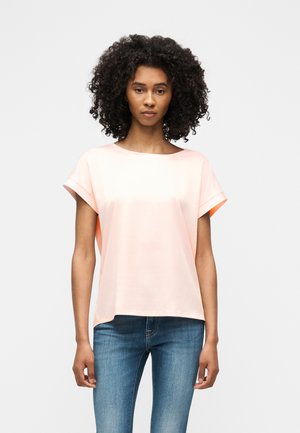 Fiatal nő göndör hajjal, világos rózsaszín rövid ujjú felsőt és kék farmert viselve, tiszta fehér háttér előtt áll.