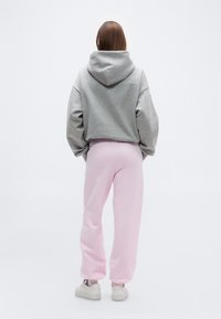 Άτομο που στέκεται γυρισμένο προς τα πίσω, φοράει γκρι φούτερ με κουκούλα, ανοιχτό ροζ φόρμα και λευκά αθλητικά παπούτσια, μπροστά σε λευκό απλό φόντο.