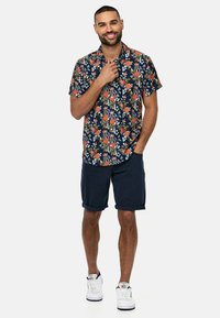 La camisa de flores de manga corta en color azul marino presenta patrones coloridos con acentos en rojo y verde, combinada con pantalones cortos azules enrollados y zapatillas blancas.